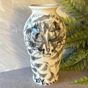 May include: A white ceramic vase with a black and gray cat face design. The vase has a narrow neck and a wider body, with the cat's features painted in a stylized, artistic manner. The vase is a decorative home decor item.