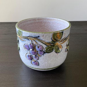 May include: A white ceramic planter with a grapevine design. The planter has a light pink interior and is decorated with green leaves, purple grapes, and gold accents.
