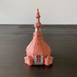 May include: A small, terracotta-colored ceramic church with a white-trimmed spire and a green-painted door. The church has a detailed roof and windows.