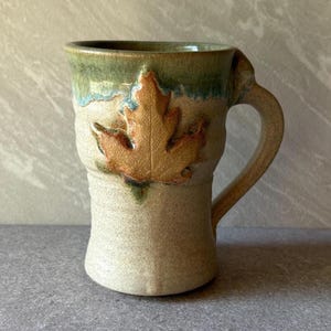 May include: A ceramic mug with a speckled cream-colored base and a textured, raised maple leaf design in shades of orange and brown. The upper rim features a smooth, glossy green glaze that drips down slightly over the textured leaf surface.
