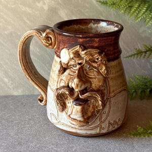 May include: A ceramic mug featuring a sculpted face with a prominent nose, mustache, and beard. The mug has a dark brown glazed rim and handle, transitioning to a lighter, speckled beige body. The name J. Duc is etched near the base.