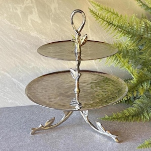 May include: A two-tiered serving stand crafted from silver-toned metal with a hammered texture on both circular trays. The central support column and the three tripod legs feature a decorative branch design with small leaf accents.