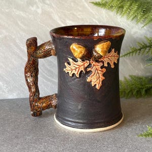 May include: A dark brown ceramic mug with a textured surface and a handle shaped like a tree branch. The exterior features a raised, three-dimensional relief of two golden-brown acorns and several light brown oak leaves attached to the side.