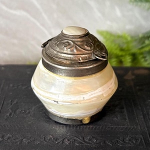 May include: An antique, pear-shaped container with a silver-toned lid and a cream-colored body. The lid features ornate detailing and a small, round, off-white stone. The body has horizontal white stripes. The container is likely used for storing small items.