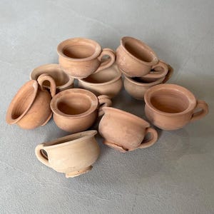 May include: A collection of small, unglazed terracotta cups with handles. The cups are various sizes and shapes, with a natural, earthy tone. The cups are scattered on a light gray surface.