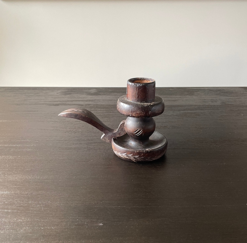 Pu&ograve; includere: Un portacandele in legno marrone scuro con un design intagliato. Il portacandele ha una piccola maniglia sul lato e una base rotonda.