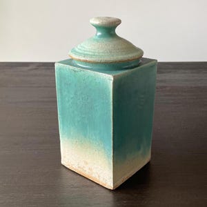 May include: A square, ceramic jar with a lid. The jar is a gradient of turquoise to off-white, with a matching lid. The lid has a small, rounded handle. The jar is sitting on a dark brown surface.