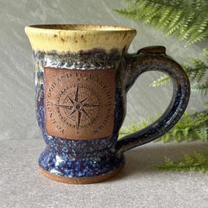 May include: A ceramic mug with a speckled blue and dark glaze on the base and a cream-colored rim. A square terracotta-colored panel on the front features an embossed compass rose design with the text TO KNOW GOD AND TO MAKE HIM KNOWN.