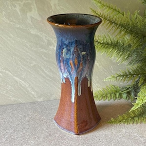 May include: A ceramic vase with a flared rim and a tapered body. The lower portion features a smooth, glossy brown glaze, while the upper section transitions into a deep blue glaze with white drip patterns cascading down the sides of the vessel.