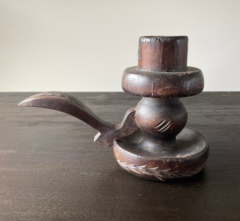 Pu&ograve; includere: Un portacandele in legno marrone scuro con un design intagliato. Il portacandele ha un manico lungo e curvo e una base rotonda.