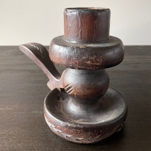 Pu&ograve; includere: Un portacandele in legno marrone scuro con un design intagliato. Il portacandele ha una base rotonda e una parte superiore cilindrica alta con una piattaforma rotonda pi&ugrave; piccola al centro. Il portacandele ha un manico sul lato.