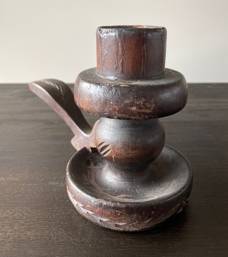 Pu&ograve; includere: Un portacandele in legno marrone scuro con un design intagliato. Il portacandele ha una base rotonda e una parte superiore cilindrica alta con una piattaforma rotonda pi&ugrave; piccola al centro. Il portacandele ha un manico sul lato.