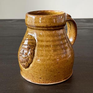 May include: A brown ceramic mug with a raised face design. The mug has a rounded body and a handle.