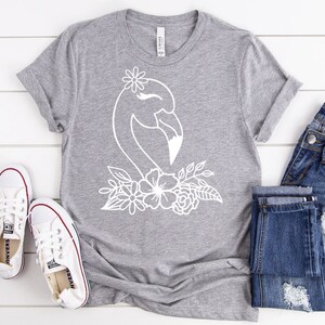 May include: A grey t-shirt with a white flamingo design and floral details. The shirt is styled with blue denim jeans and white canvas trainers with red trim. The items are on a white wooden surface.