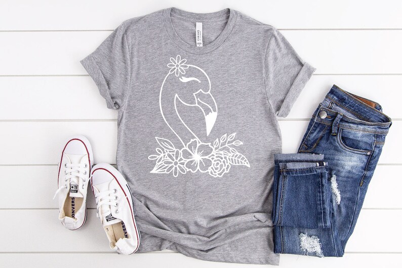 May include: A grey t-shirt with a white flamingo design and floral details. The shirt is styled with blue denim jeans and white canvas trainers with red trim. The items are on a white wooden surface.