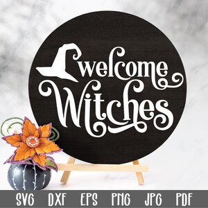 May include: A black circle sign with white text that reads "Welcome Witches" with a witch's hat silhouette above the text. The sign is on a small wooden easel.
