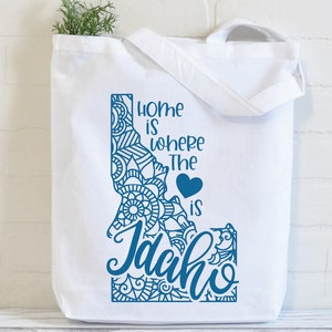 May include: White tote bag with a blue outline of the state of Idaho. The outline is filled with a blue mandala design. The text "Home is where the heart is Idaho" is written in blue cursive lettering.