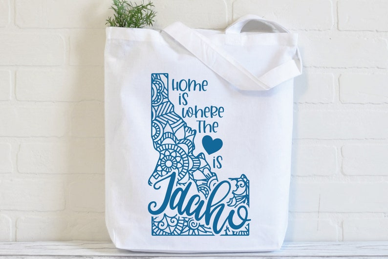 May include: White tote bag with a blue outline of the state of Idaho. The outline is filled with a blue mandala design. The text "Home is where the heart is Idaho" is written in blue cursive lettering.