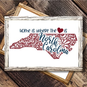 May include: A white framed sign with the text "Home is where the heart is" and a red heart symbol. The outline of the state of North Carolina is filled with a red mandala design. The words "North Carolina" are written in blue inside the state outline.