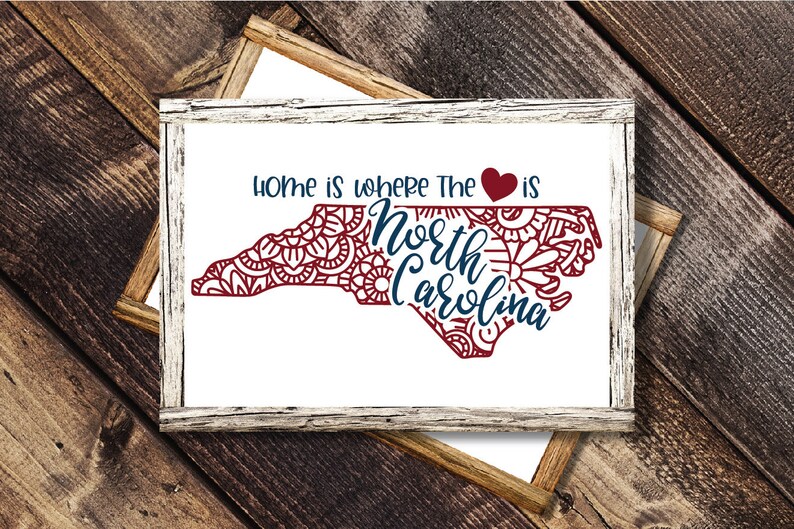 May include: A white framed sign with the text "Home is where the heart is" and a red heart symbol. The outline of the state of North Carolina is filled with a red mandala design. The words "North Carolina" are written in blue inside the state outline.