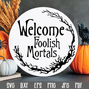 May include: A white round sign with black lettering that reads "Welcome Foolish Mortals" surrounded by a black twig wreath. The sign is sitting on a table with two pumpkins and a black crow.
