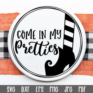 May include: A white round sign with black text and a black witch's shoe. The text reads "Come in my pretties".