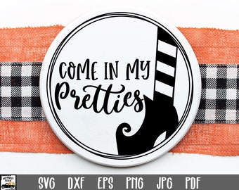 May include: A white round sign with black text and a black witch's shoe. The text reads "Come in my pretties".