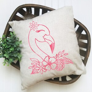 May include: A square, beige cushion with a pink flamingo design. The flamingo has a flower crown and is surrounded by pink flowers and leaves. The cushion is placed in a dark brown wooden basket.