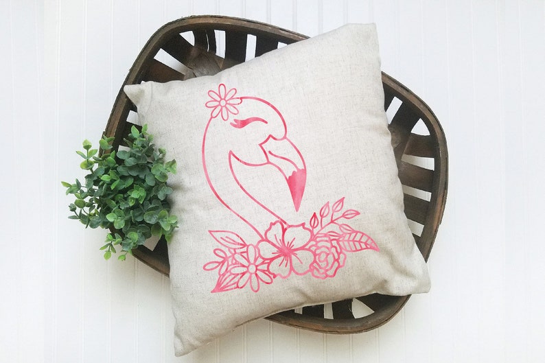 May include: A square, beige cushion with a pink flamingo design. The flamingo has a flower crown and is surrounded by pink flowers and leaves. The cushion is placed in a dark brown wooden basket.
