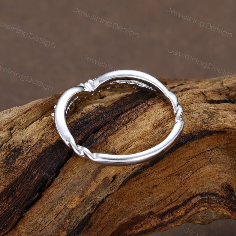 Pu&ograve; includere: Un anello d'argento con un design a fascia intrecciata. L'anello presenta una fila di piccole pietre trasparenti lungo la curva superiore. L'anello &egrave; posto su uno sfondo di legno, evidenziando il suo design elegante. &Egrave; un gioiello.