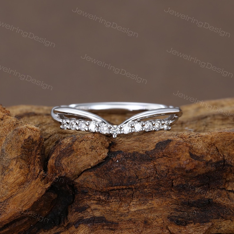 Pu&ograve; includere: Un anello argentato curvo con una fila di piccole pietre preziose trasparenti. L'anello &egrave; appoggiato su una superficie di legno marrone strutturata. Le parole "Jewelring Design" sono visibili sullo sfondo.