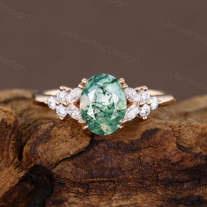 Puede incluir: Un anillo de oro con una piedra preciosa de ágata musgosa verde de corte ovalado. El anillo está flanqueado por piedras preciosas blancas en forma de marquesa a cada lado. El anillo está sobre una superficie de madera.