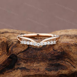Pu&ograve; includere: Un anello in oro rosa con un design curvo, incastonato con piccole pietre preziose trasparenti. L'anello &egrave; esposto su una superficie di legno strutturata, che mostra i suoi delicati dettagli e la sua maestria. Il design dell'anello &egrave; elegante e moderno.