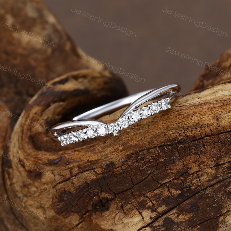 Pu&ograve; includere: Un anello color argento con un design curvo. L'anello presenta una fila di piccole pietre preziose trasparenti lungo la curva. L'anello &egrave; esposto su un pezzo di legno marrone strutturato.