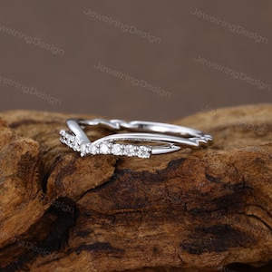 Pu&ograve; includere: Un anello color argento con un design a forma di V, ornato da piccole pietre preziose scintillanti. L'anello &egrave; posto su una superficie di legno marrone scuro strutturata, che ne mette in risalto gli eleganti dettagli. &Egrave; un gioiello.