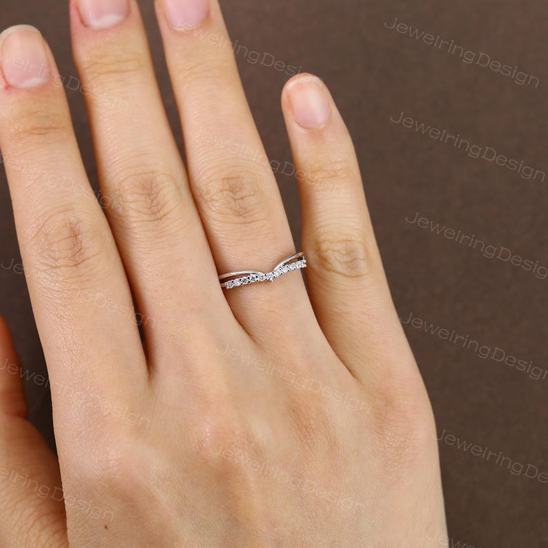 Pu&ograve; includere: Un anello color argento con un design curvo, incastonato con piccole pietre preziose trasparenti. L'anello &egrave; mostrato sulla mano di una persona, con uno sfondo neutro. Il design dell'anello &egrave; a forma di V, con le gemme incastonate lungo la curva superiore.