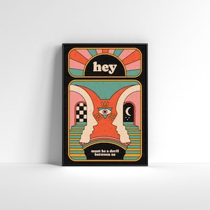 Op de afbeelding: Een ingelijste kunstdruk met een retro-ontwerp. Het kunstwerk toont het woord "hey" boven een zonnestraal en twee tegenover elkaar liggende silhouetten met een oogsymbool. De tekst "must be a devil between us" staat onderaan. Het kleurenpalet omvat oranje, roze, teal en zwart.