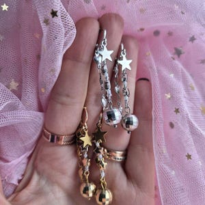 May include: Three pairs of dangling earrings are displayed. Two pairs are silver with star and disco ball charms. The third pair is gold with star and ball charms. The background is pink tulle with gold stars.