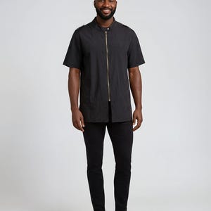 Op de afbeelding: Een persoon draagt een zwart shirt met korte mouwen met een gouden rits en een zwarte broek. Het shirt heeft een opstaande kraag en epauletten. De persoon draagt ook grijze sneakers.