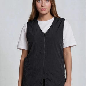 May include: A black vest with a V-neck and a full-length zipper. The vest has two front pockets and is worn over a white t-shirt. The model is wearing black pants. The vest appears to be made of a crinkled fabric.
