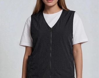 Può includere: Un gilet nero con scollo a V e cerniera a tutta lunghezza. Il gilet ha due tasche frontali e viene indossato sopra una maglietta bianca. La modella indossa pantaloni neri. Il gilet sembra essere realizzato in un tessuto stropicciato.