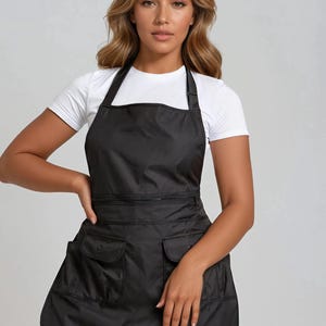 Könnte beinhalten: Schwarze Schürze mit Latz und Hüfttaschen. Die Schürze hat eine Reißverschlusstasche über der Taille und zwei quadratische Taschen auf der unteren Hälfte. Die Schürze wird über einem weißen T-Shirt und schwarzen Leggings getragen.