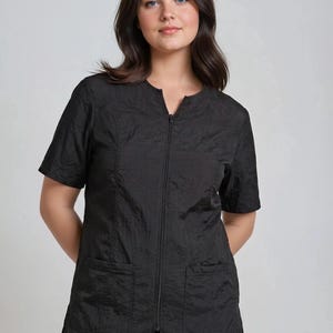 May include: Black short-sleeve zip-up top with two front pockets. The top has a textured, crinkled appearance. The model is wearing black leggings. The top is a solid black color.