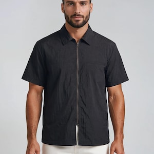 May include: Black short-sleeve shirt with a full-length zipper closure. The shirt has a collared neckline and a textured appearance. The model is wearing the shirt with white pants. The shirt is a solid color.