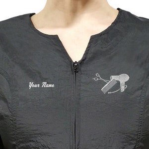 May include: Black salon smock with a zipper closure and a white embroidered design of a hair dryer, comb, and shears. The text "Your Name" is also embroidered on the smock.