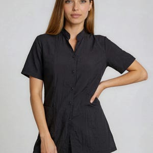 Könnte beinhalten: Schwarze kurzärmlige Tunika mit Stehkragen und Knopfleiste. Die Tunika hat eine strukturierte Optik und eine einzelne Tasche auf der linken Seite. Das Model trägt schwarze Leggings.