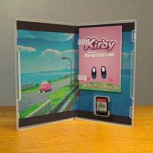 May include: An open video game case with a pink instruction manual that says "Kirby and the Forgotten Land" and a game cartridge. The case has a blue and green background with a car driving on a road.