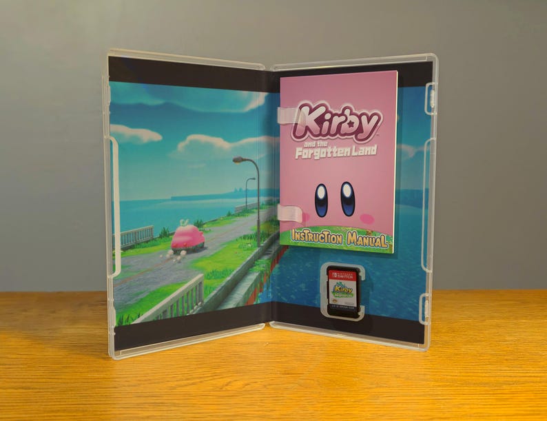 May include: An open video game case with a pink instruction manual that says "Kirby and the Forgotten Land" and a game cartridge. The case has a blue and green background with a car driving on a road.