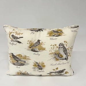 May include: A white pillow with a brown and gold bird print. The pillow features different types of birds, including a bullfinch, a starling, a rook, a kingfisher, a whinchat, a jay, a wren, and a short-eared owl. The names of the birds are printed on the pillow.