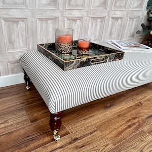 May include: A black and white striped ottoman with wooden legs and brass castors. A black tray with a marble design sits on top of the ottoman, holding two glass candle holders with orange candles.
